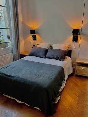 A neatly made bed with a dark blanket, two bedside tables with lamps, a window with curtains, and a radiator. -     Parisian Elegance & Modern Comfort on Île Saint-Louis (Paris)