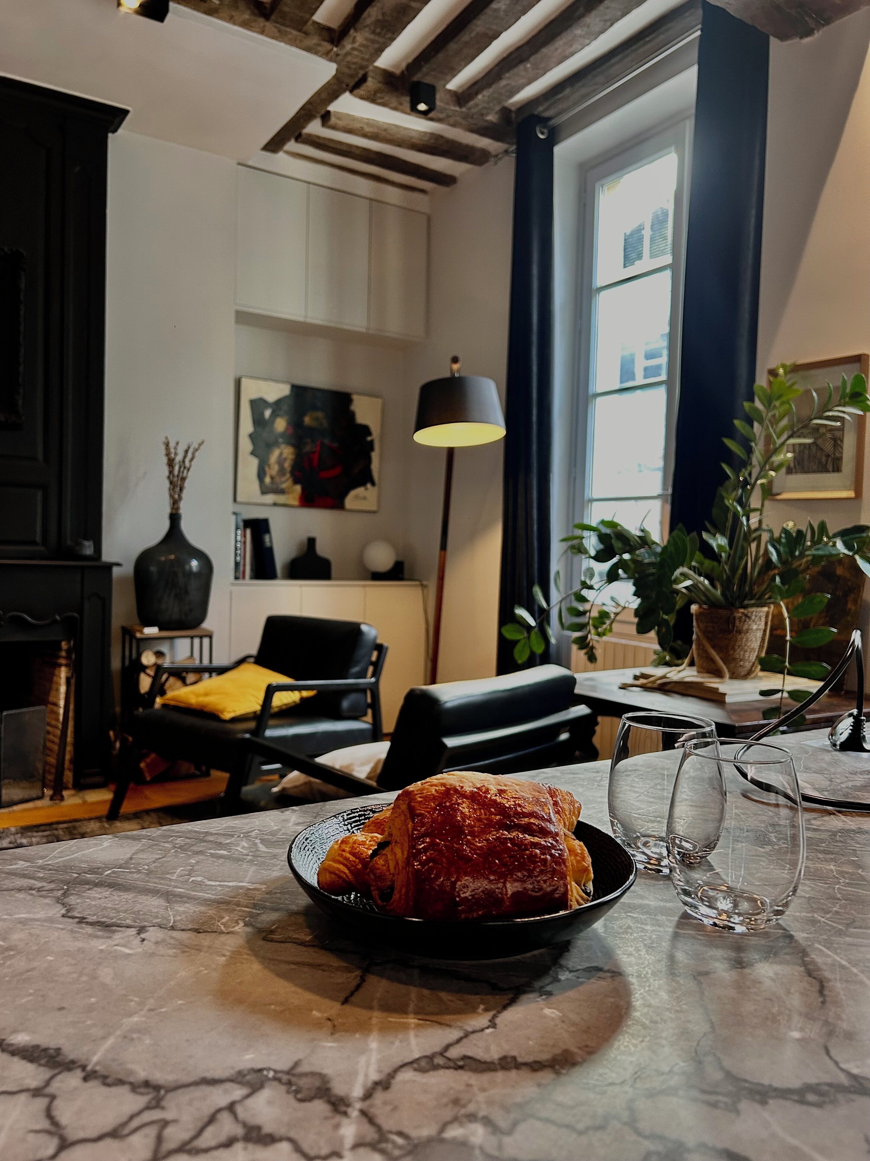 Un salon chaleureux avec une cheminée, des fauteuils confortables, une table à manger avec une assiette de nourriture et une grande fenêtre avec des rideaux.