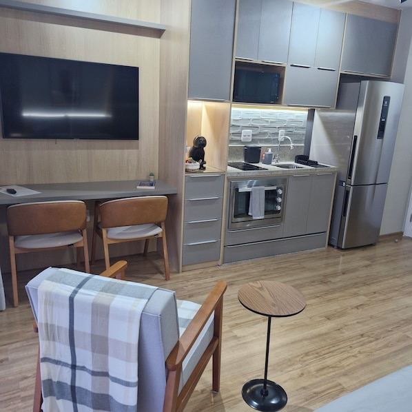 A modern kitchen with a flat-screen TV, a dining area with two chairs, a small table, and a sofa with a checkered cover. - Fully equipped studio apartment with queen bed, 140m from Alto do Ipiranga metro station, with air conditioning. (São Paulo)