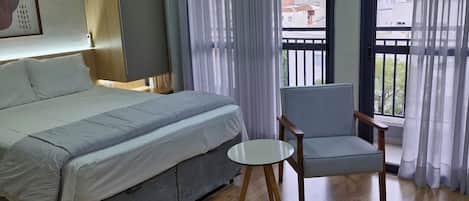 A hotel room with a bed, a chair, a small table, and a window with curtains.