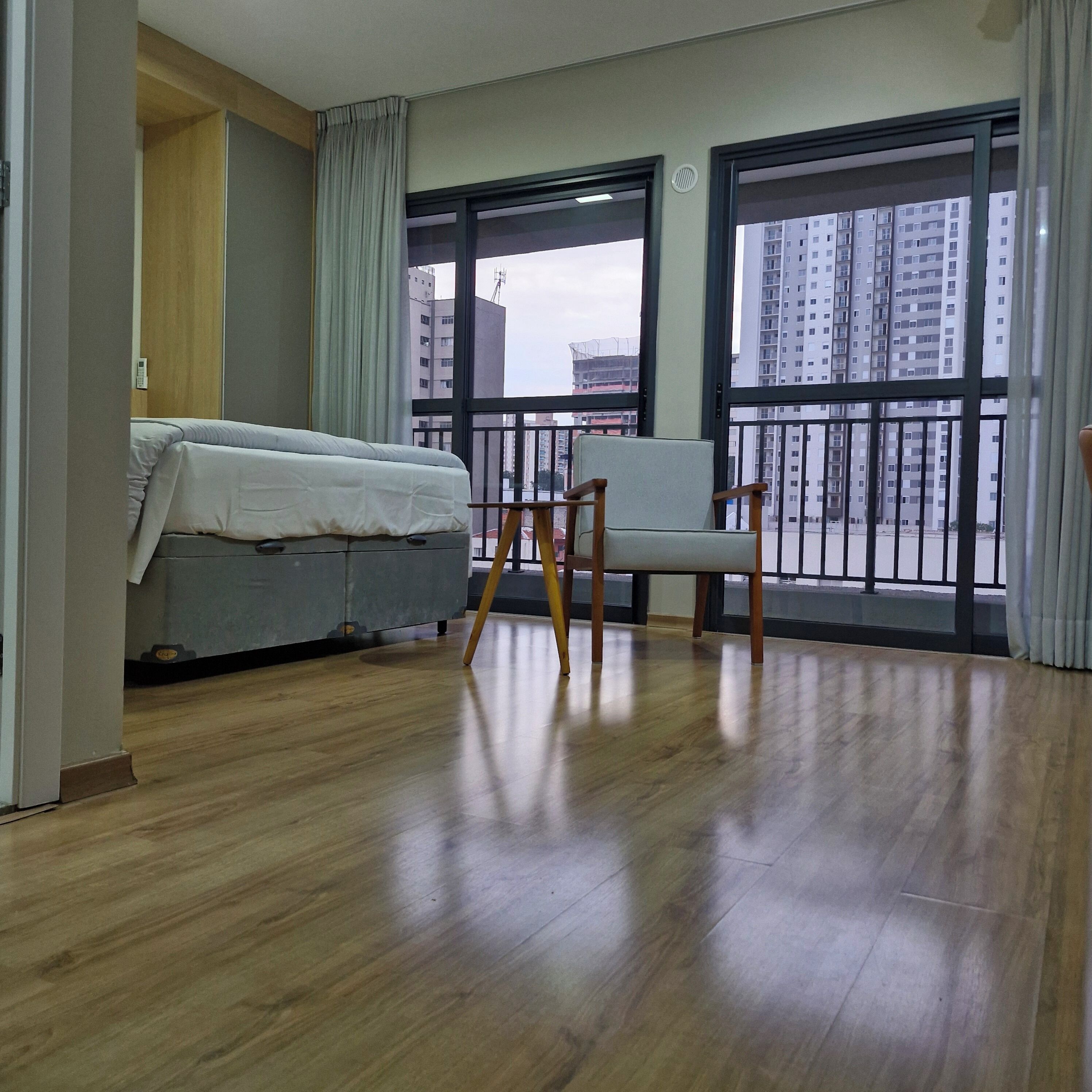 Quarto de hotel com cama, uma cadeira, janela com vista para edifícios altos e piso de madeira.