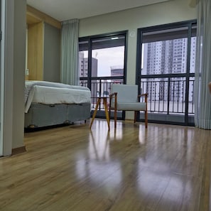 A hotel room with a bed, a chair, a window with a view of high-rise buildings, and a wooden floor. - Fully equipped studio apartment with queen bed, 140m from Alto do Ipiranga metro station, with air conditioning. (São Paulo)