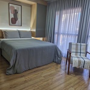 Quarto de hotel com cama, uma cadeira, uma janela grande com cortinas e uma obra de arte na parede.