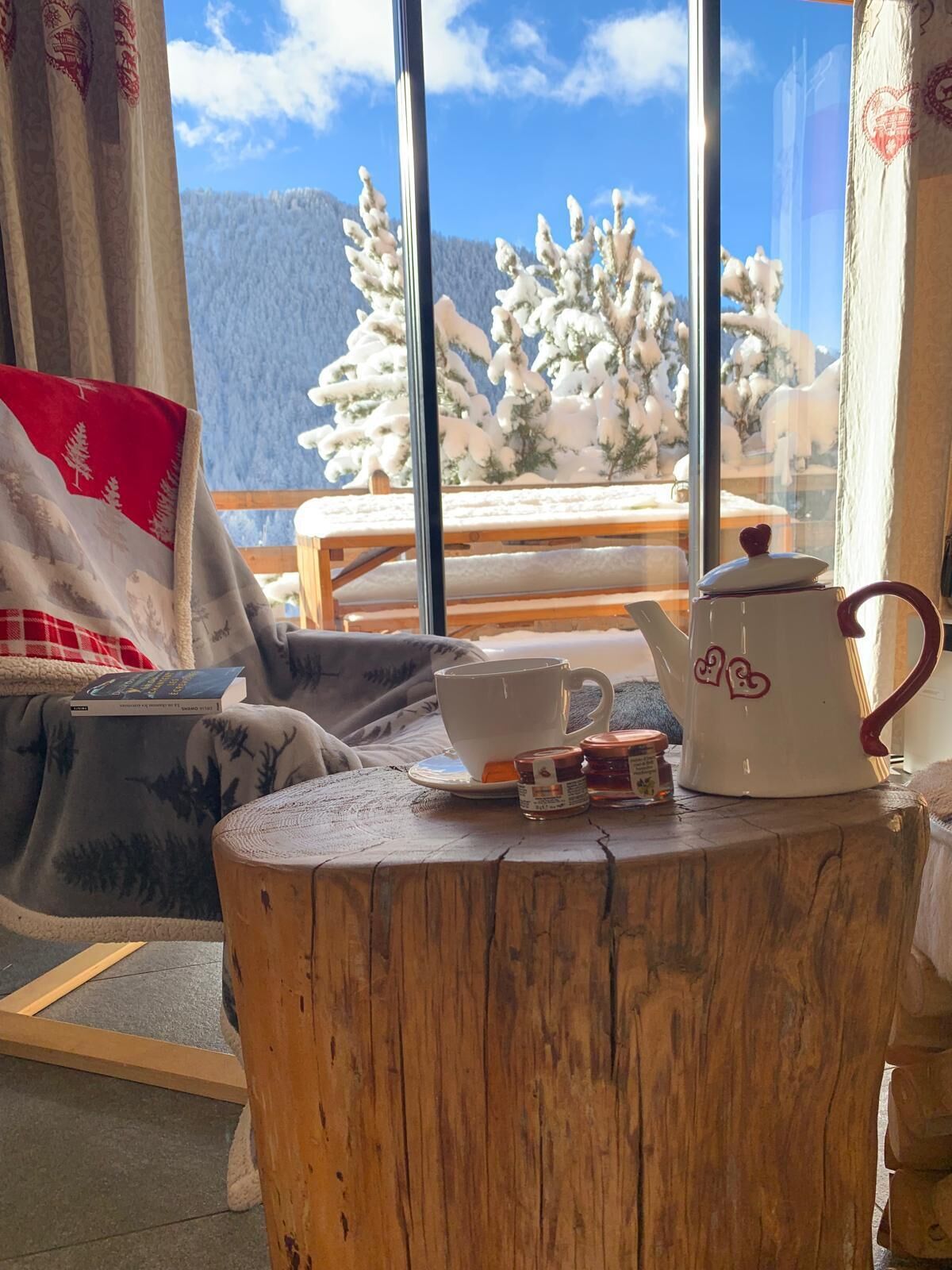 Ein gemütliches Zimmer mit Blick auf schneebedeckte Bäume, ein Holztisch mit Teekanne, Tasse und Einmachglas sowie ein Stuhl mit Decke.