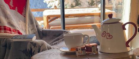 Ein gemütliches Zimmer mit Blick auf schneebedeckte Bäume, ein Holztisch mit Teekanne, Tasse und Einmachglas sowie ein Stuhl mit Decke.
