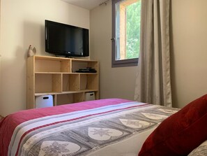 A bedroom with a bed, a television on a wooden shelf, and a window with curtains. - Vrbo Property (Molines-en-Queyras)