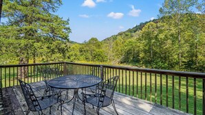 A wooden deck with a round table and chairs, surrounded by a metal railing, overlooking a forested area. - South Fork River Farmhouse with River Access (Stanton)
