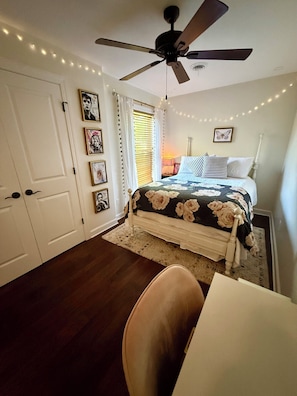 Ein Schlafzimmer mit einem Bett, einem Deckenventilator, einem Fenster mit Jalousien und Lichterketten.