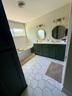Une salle de bain avec une baignoire, un lavabo, des meubles verts, un grand miroir et un sol carrelé à motifs hexagonaux.