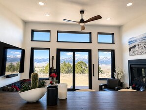 A spacious room with a large dining table, multiple windows offering mountain views, a ceiling fan, and a fireplace. - Modern Mountain Escape 2 Bed, 2 Bath 360 degree Sangre Views (Westcliffe)