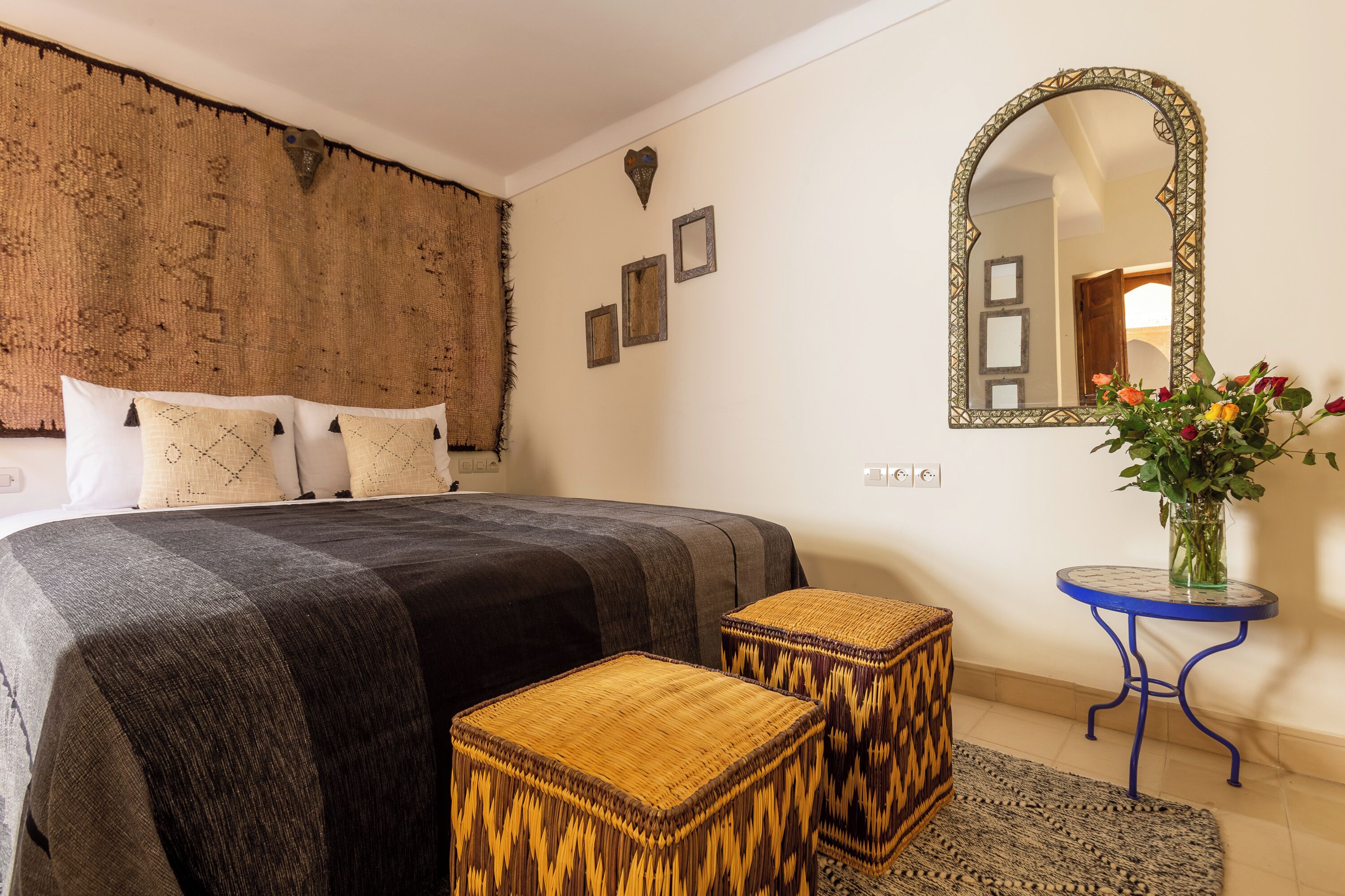 A bedroom with a bed, a mirror, a small table with a vase of flowers, and a woven stool.