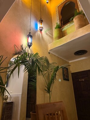 Une pièce avec un plafond haut, une chaise en bois, une plante en pot, une étagère fixée au mur avec une plante, et une porte.
