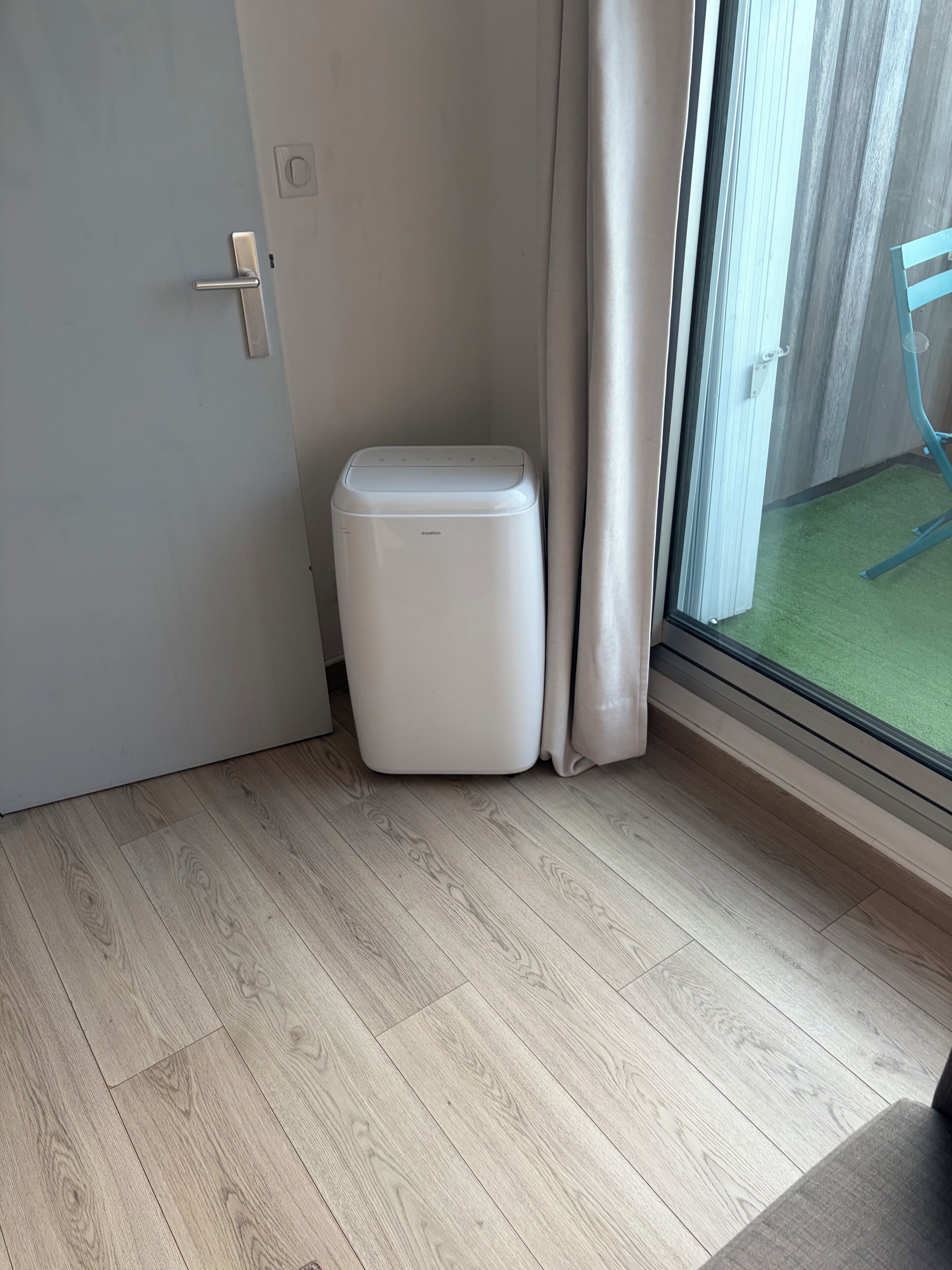A room with a wooden floor, a white trash bin, a door, and a window with a view of a green outdoor area.