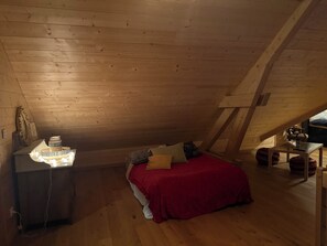 A small, cozy attic bedroom with a sloped ceiling, a bed with a red blanket, a bedside table with a lamp, and a wooden dresser.