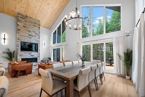 A spacious dining area with a large wooden table and modern chairs, a stone fireplace, and large windows offering a view of the outdoors.