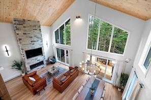 A spacious living room with a stone fireplace, large windows, and a wooden ceiling.