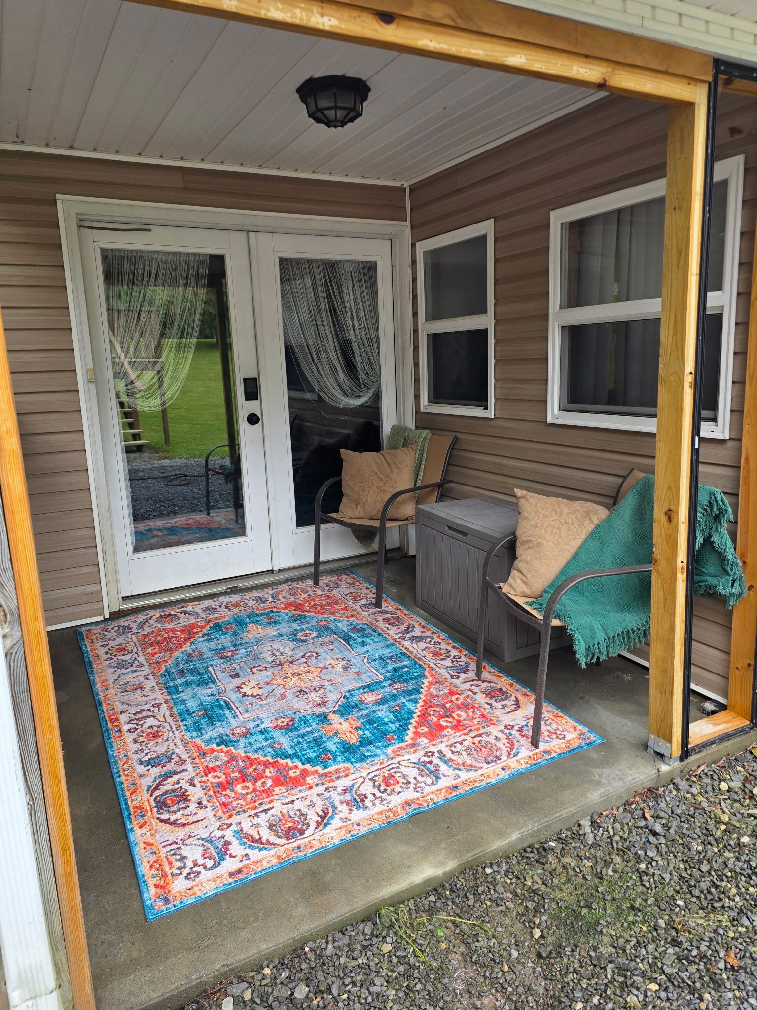 A covered porch with a patterned rug, a bench, and a door with curtains.