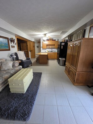 A living room with a sofa, a coffee table, and a rug. There is a kitchen area with wooden cabinets and a refrigerator.