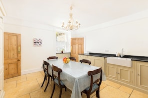 A dining area with a table set for four, a chandelier, and a kitchen with a sink and cabinets. - The Village Farmhouse | Cotswolds | Sleeps 6 - Pass the Keys (Cirencester)