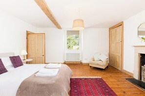 A bedroom with a bed, a chair, a fireplace, and a window with a view of greenery. - The Village Farmhouse | Cotswolds | Sleeps 6 - Pass the Keys (Cirencester)