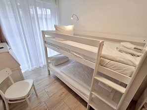 Uma cama beliche com escrivaninha e cadeira, uma janela com cortinas e uma luminária fixada na parede.