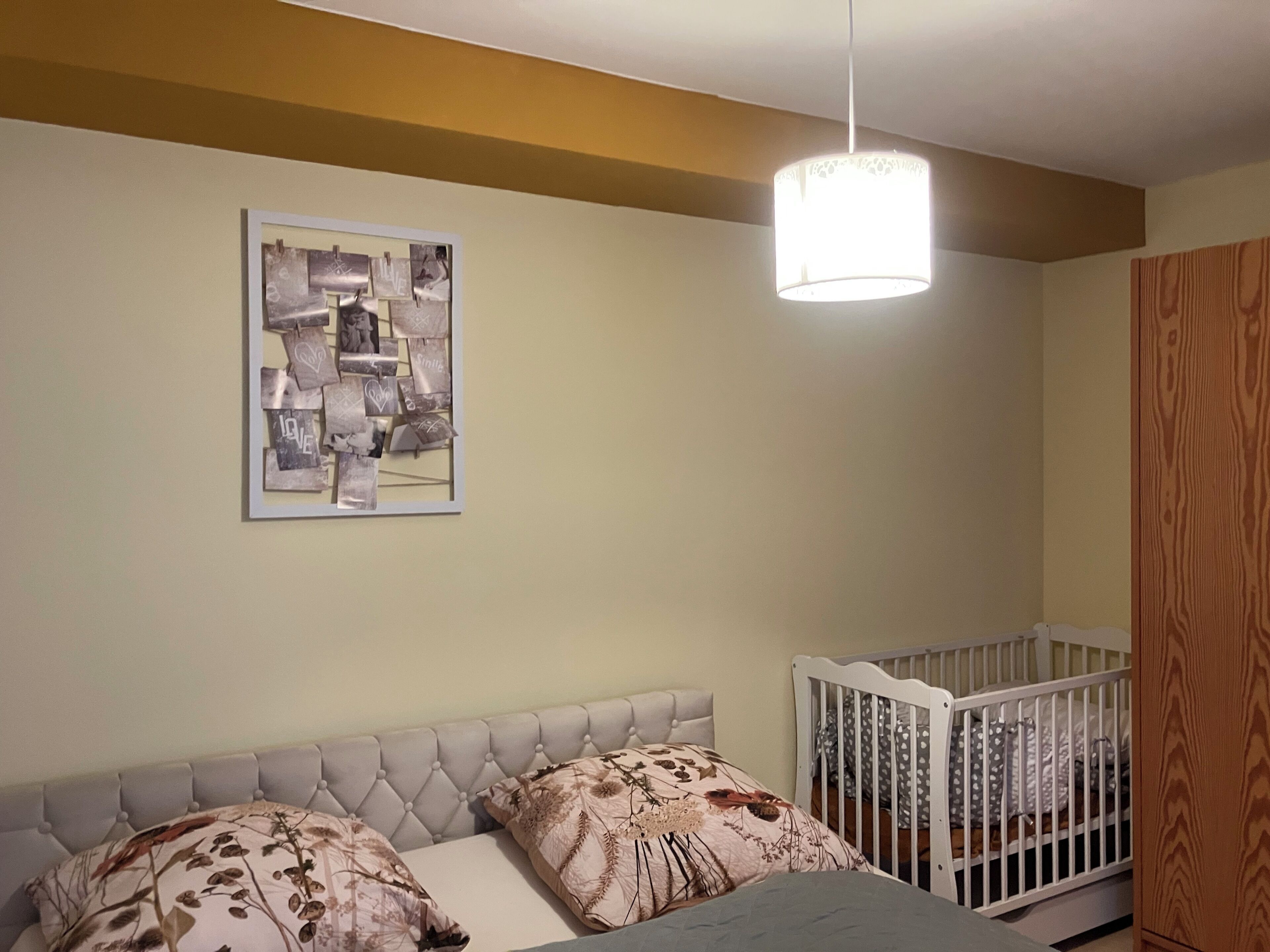 Ein Schlafzimmer mit einem Bett, einem Babybett, einer hängenden Deckenleuchte und einem gerahmten abstrakten Kunstwerk an der Wand.