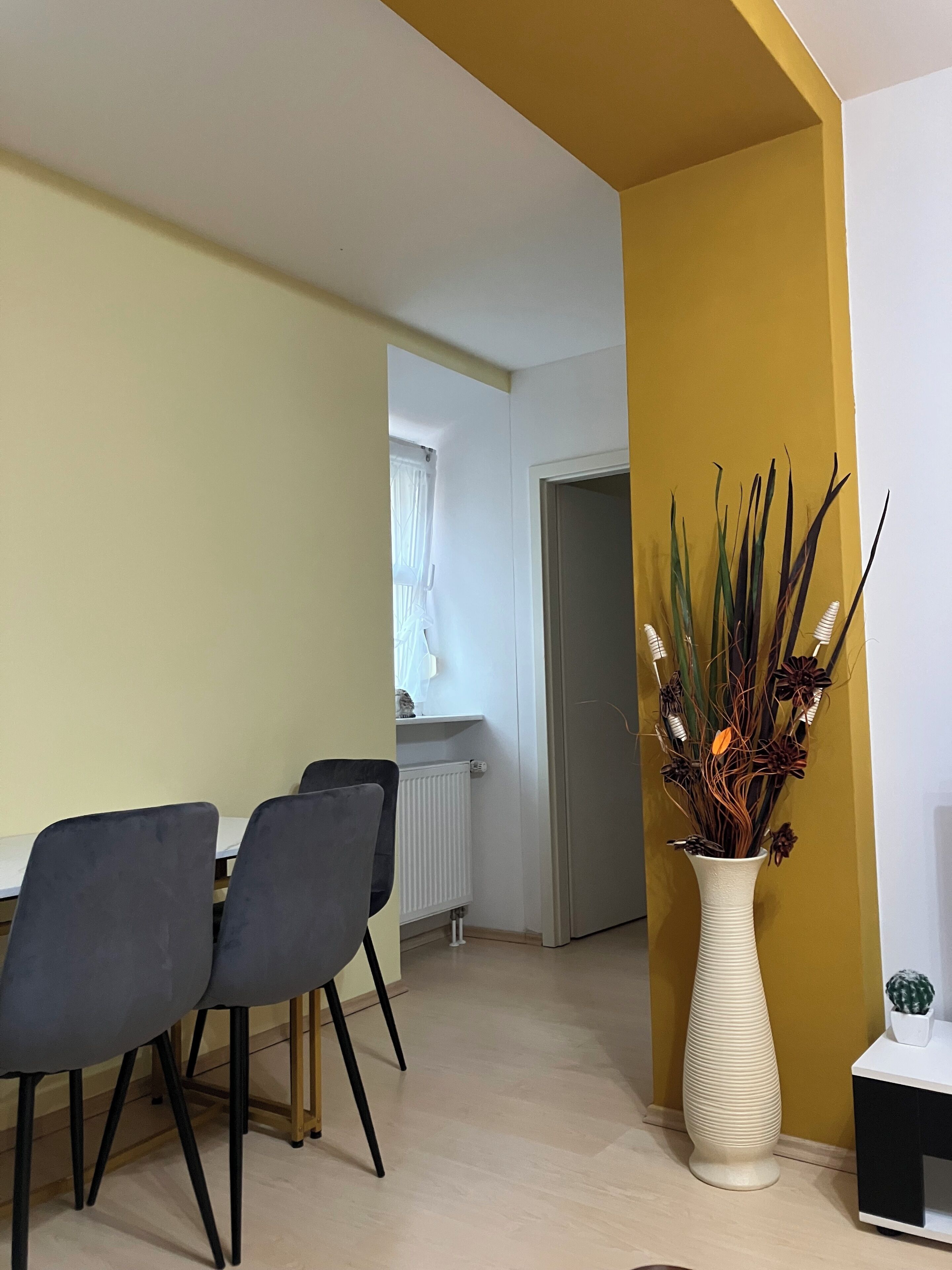 Un comedor con una mesa y sillas, un jarrón con plantas, y una puerta que da a otra habitación.