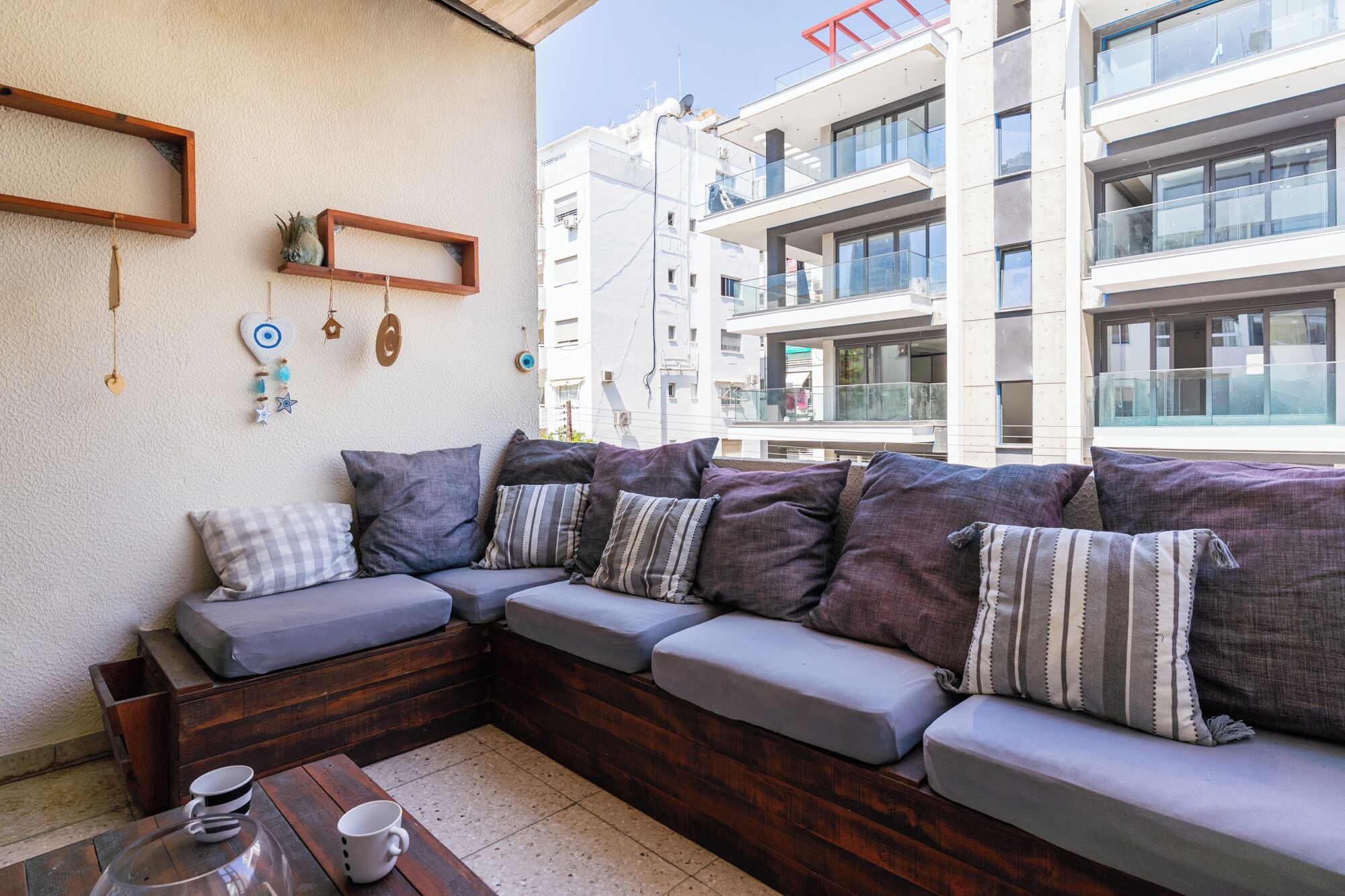 Uma área de estar moderna ao ar livre, com um banco de madeira e almofadas, uma mesa de centro com xícaras e uma parede com objetos decorativos.