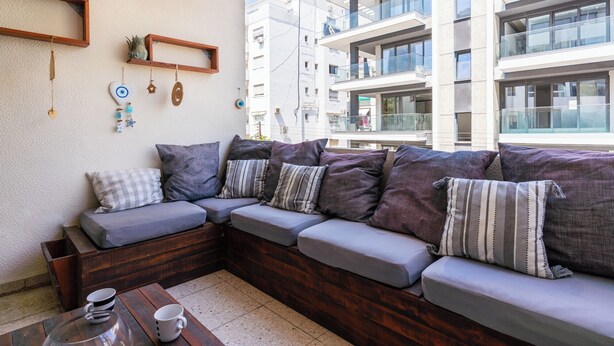 Un espacio exterior moderno con una banca de madera y cojines, una mesa de centro con tazas, y una pared con adornos.