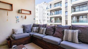 Uma área de estar moderna ao ar livre, com um banco de madeira e almofadas, uma mesa de centro com xícaras e uma parede com objetos decorativos.
