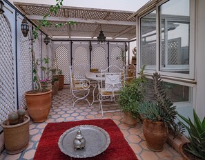 Een overdekt terras met een tafel, stoelen, potplanten en een decoratieve metalen dienblad.