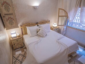 Een net opgemaakt bed met witte lakens, een bedlampje en een decoratieve wandversiering.