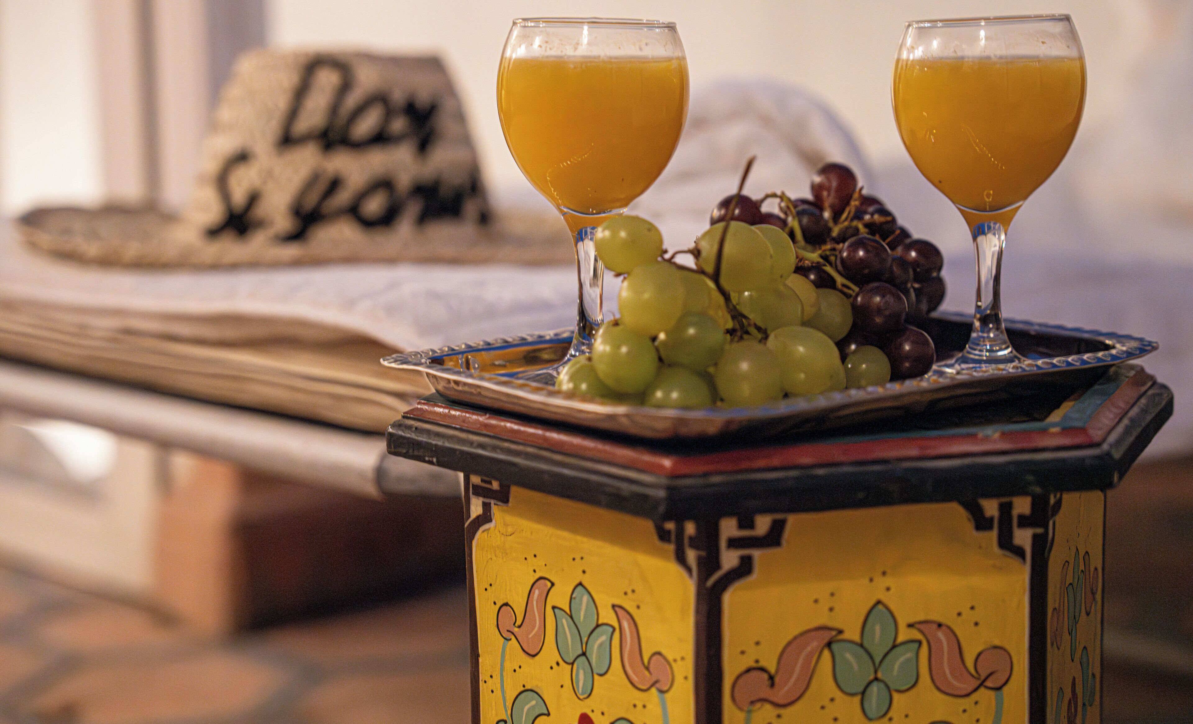 A table with two glasses of orange juice, a plate of grapes, and a decorative hat with text.