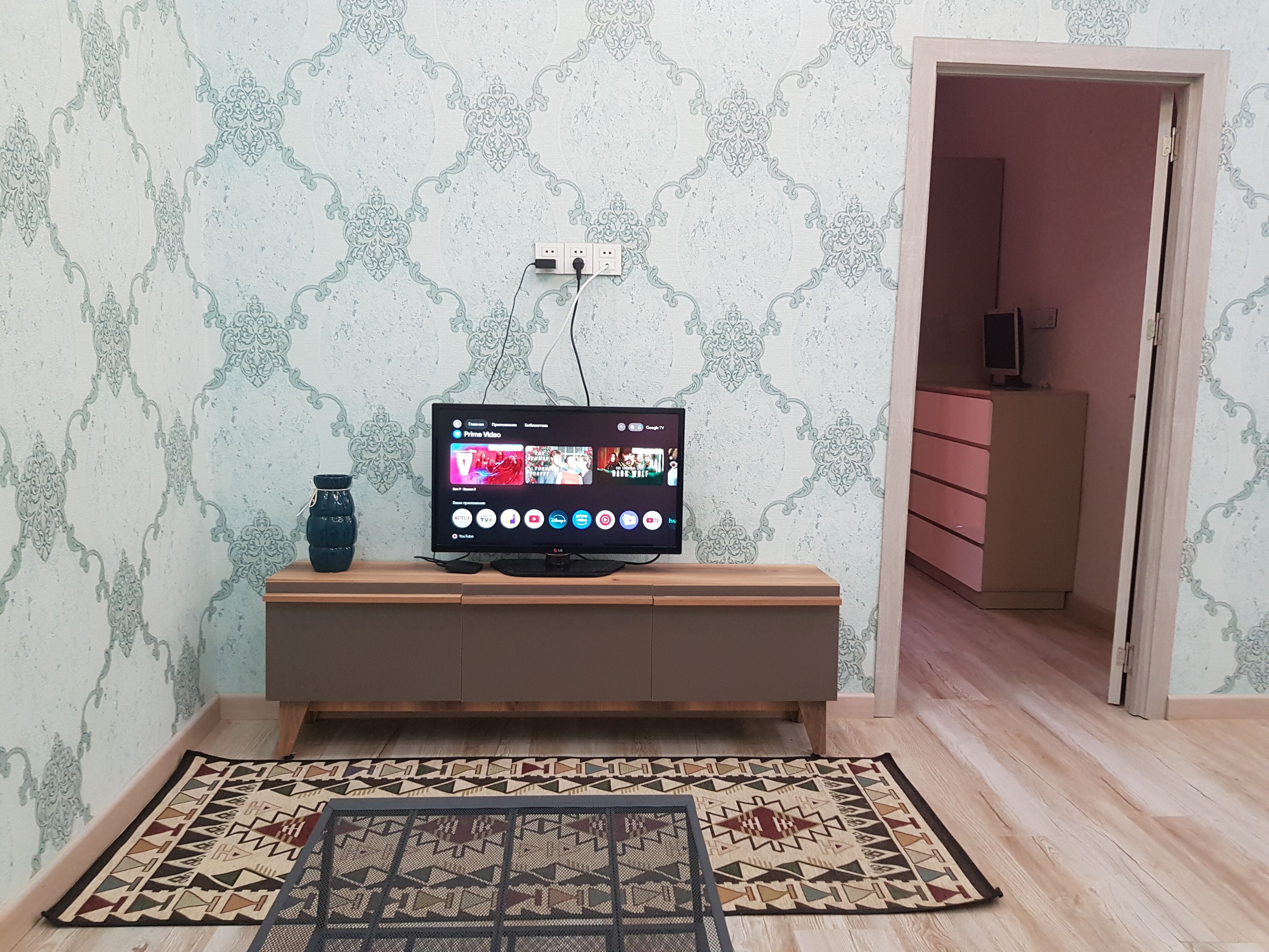 Un téléviseur posé sur un meuble en bois, un tapis à motifs et une porte-fenêtre donnant sur une autre pièce.