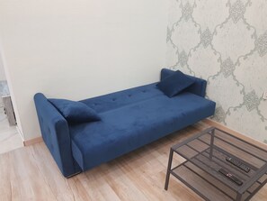 A room with a blue sofa against a wall with a patterned design, a small table with two remotes, and wooden flooring. - Cozy 1-bedroom apartment in the Baku center (Bakı)