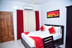 Habitación de hotel con puerta de madera, cama con ropa blanca y detalles rojos, ventilador de techo y aire acondicionado de pared.