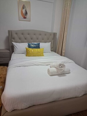 Een net opgemaakt bed met witte beddengoed, een grijs bekleed hoofdbord en een geel kussen met tekst.