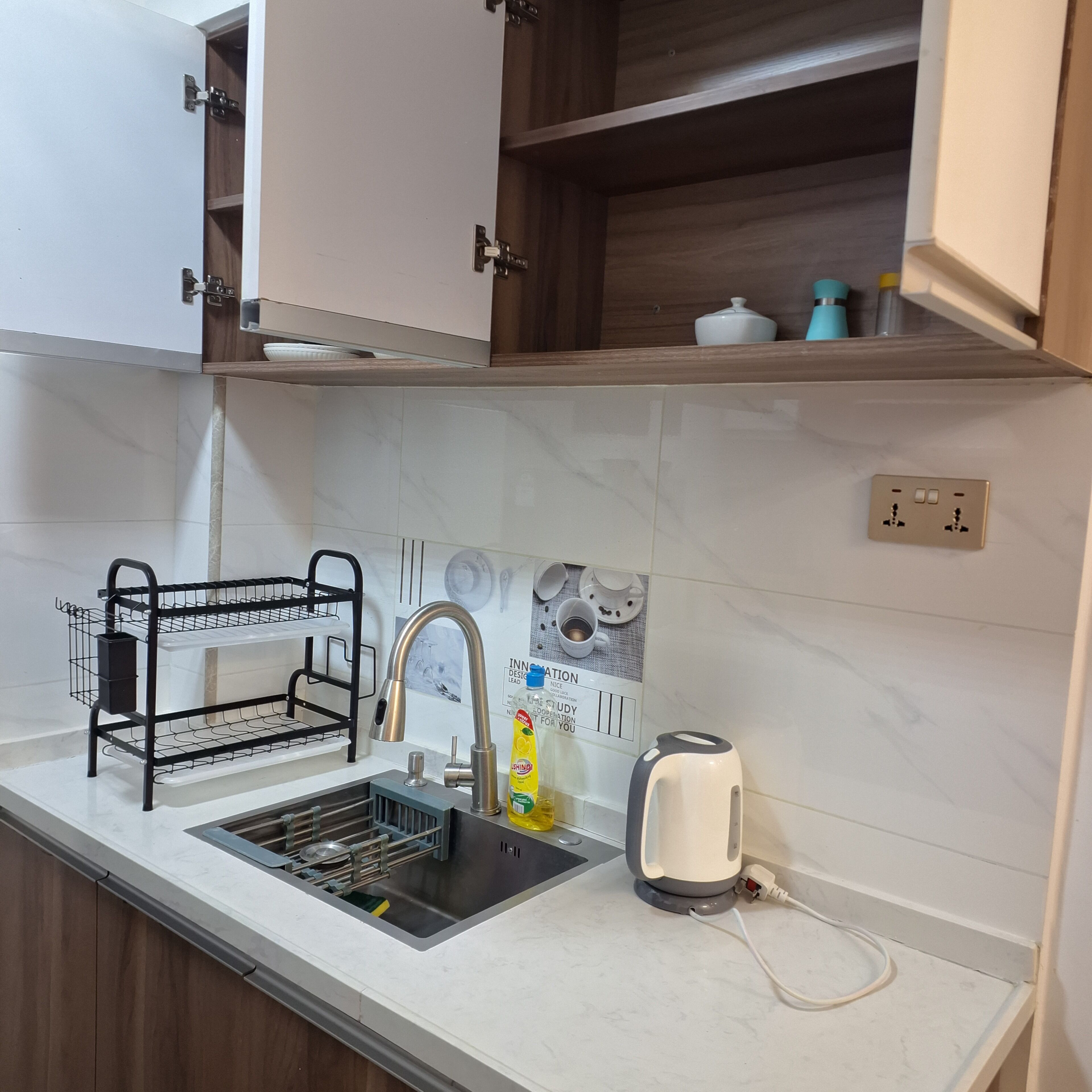 A kitchen with a sink, a black wire rack, a kettle, and a wall-mounted electrical outlet.