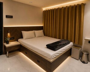 A modern bedroom with a bed, bedside tables, a nightstand, a trash bin, and a wall-mounted light fixture. - Sai Presenta Homestay (Hospet)