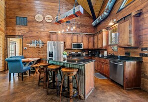 A rustic kitchen with wooden cabinets, a stainless steel refrigerator, a microwave, and a bar area with stools. - Game Loft | Hot Tub | Fire Pit | Boat Parking (Broken Bow)