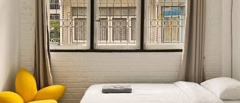 Una habitación de hotel con dos camas, una ventana con rejas decorativas de hierro, una silla amarilla y un equipo de aire acondicionado.