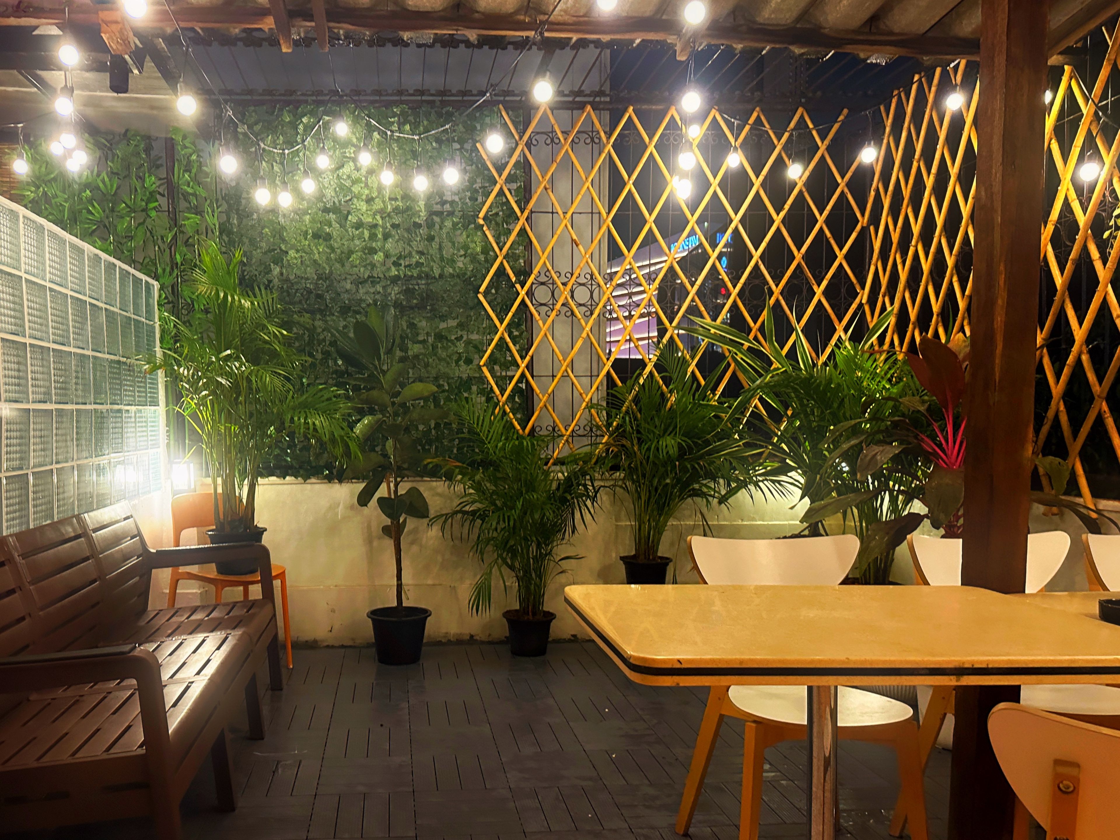 Een terras met houten banken en tafels, omringd door potplanten en lichtslingers.