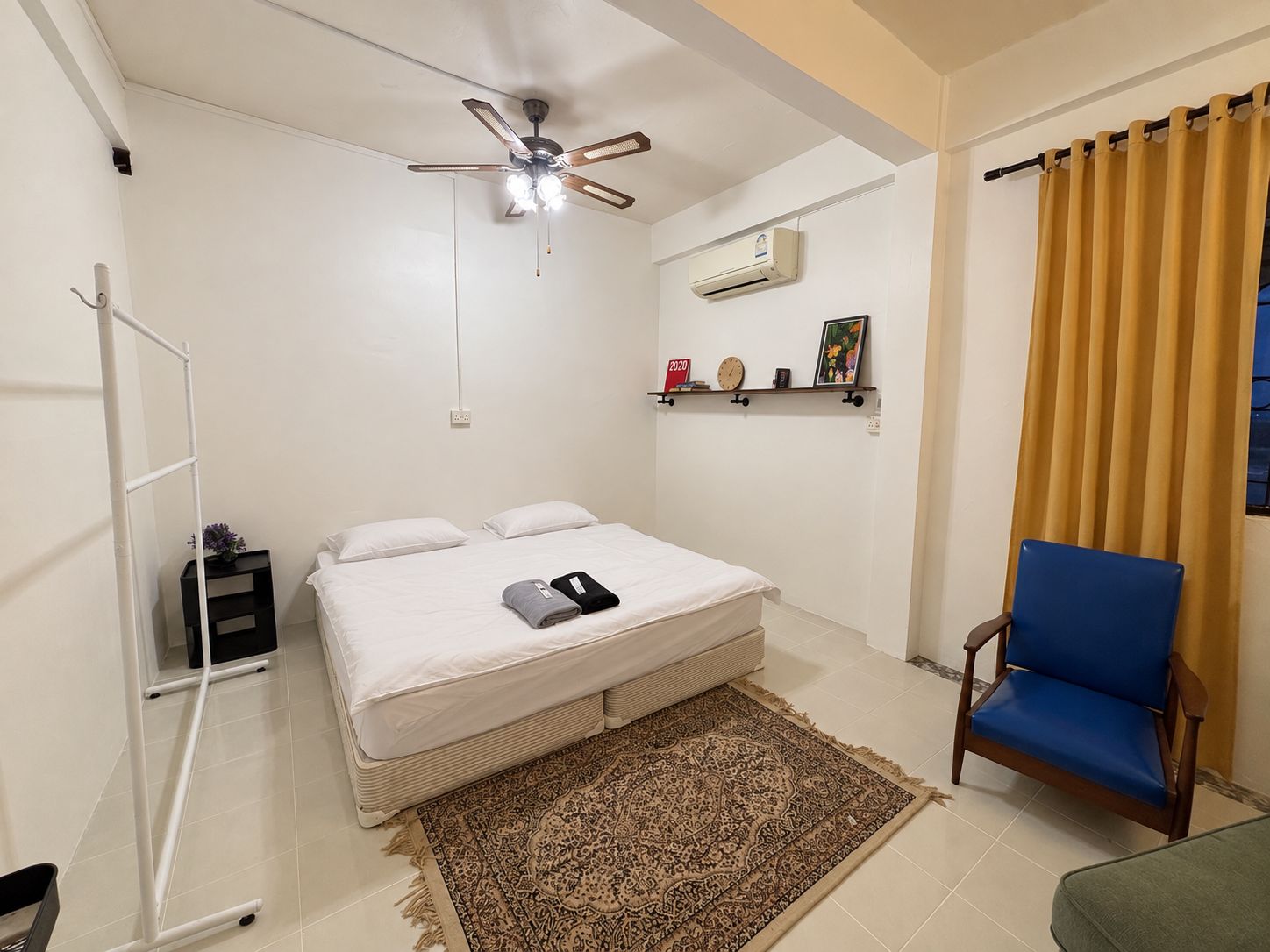 Een slaapkamer met een bed, een plafondventilator, een airconditioning, een plank met spullen, een stoel en een raam met gordijnen.