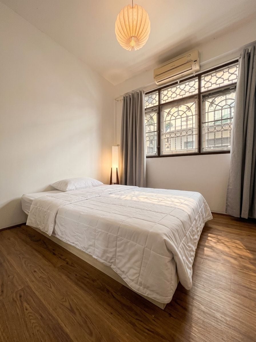 A bedroom with a bed, wooden flooring, a window with curtains, and a hanging light fixture.