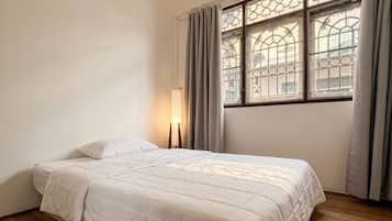 A bedroom with a bed, wooden flooring, a window with curtains, and a hanging light fixture.