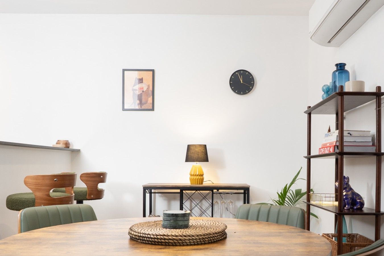 A dining area with a round wooden table, chairs, a wall-mounted clock, a framed picture, and a bookshelf with books and decorative items.