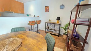 A compact living space with a dining area featuring a round wooden table and green chairs, a small kitchen with wooden cabinets, a wall-mounted clock, and a shelf with various items. - Eclectic 1BR With Private Terrace Near Manuka (Forrest)