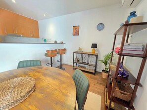 Ein kompakter Wohnraum mit Essbereich, inklusive eines runden Holztisches und grüner Stühle, einer kleinen Küche mit Holzschränken, einer an der Wand befestigten Uhr und einem Regal mit verschiedenen Gegenständen.