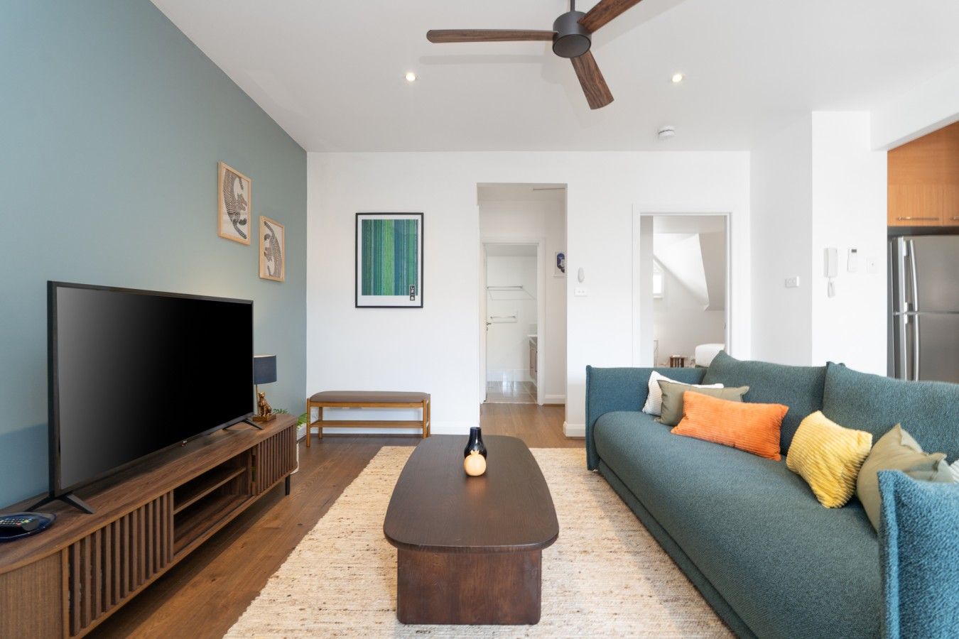 A living room with a flat-screen TV, a wooden coffee table, a sofa with cushions, a ceiling fan, and a framed artwork on the wall.