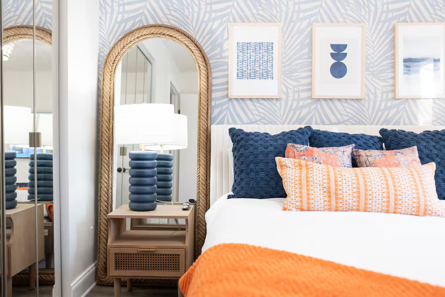 A hotel room with a large mirror, a bedside table with a lamp, a bed with multiple pillows, and framed artwork on the wall.